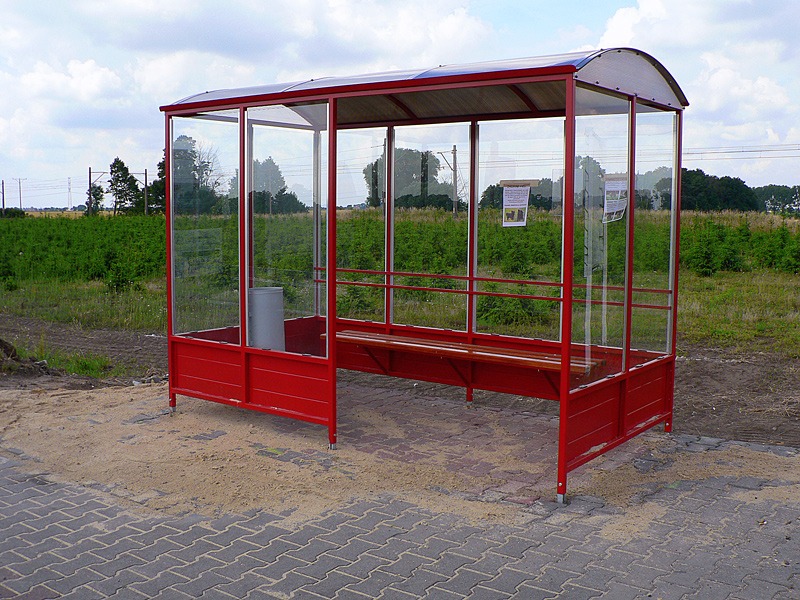 Czerwona wiata przystankowa z przezroczystymi ścianami, drewnianą ławką i koszem na śmieci, umieszczona na placu z kostki brukowej, z widoczną zielenią w tle.