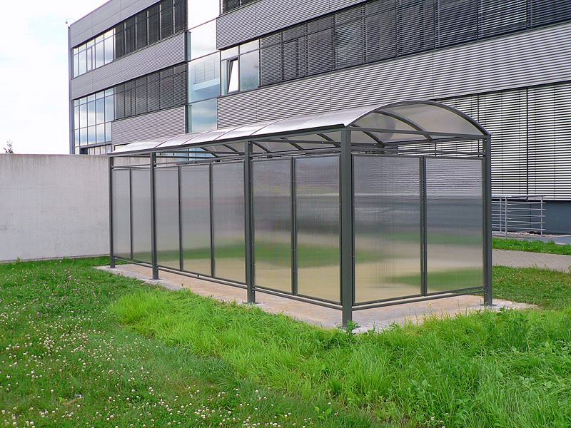 Zadaszona wiata rowerowa z przezroczystymi ścianami, umieszczona na trawniku przed nowoczesnym budynkiem biurowym.