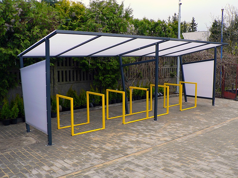 Zadaszona wiata rowerowa z żółtymi stojakami na rowery, osłonięta z boków panelami z poliwęglanu, na tle zieleni.