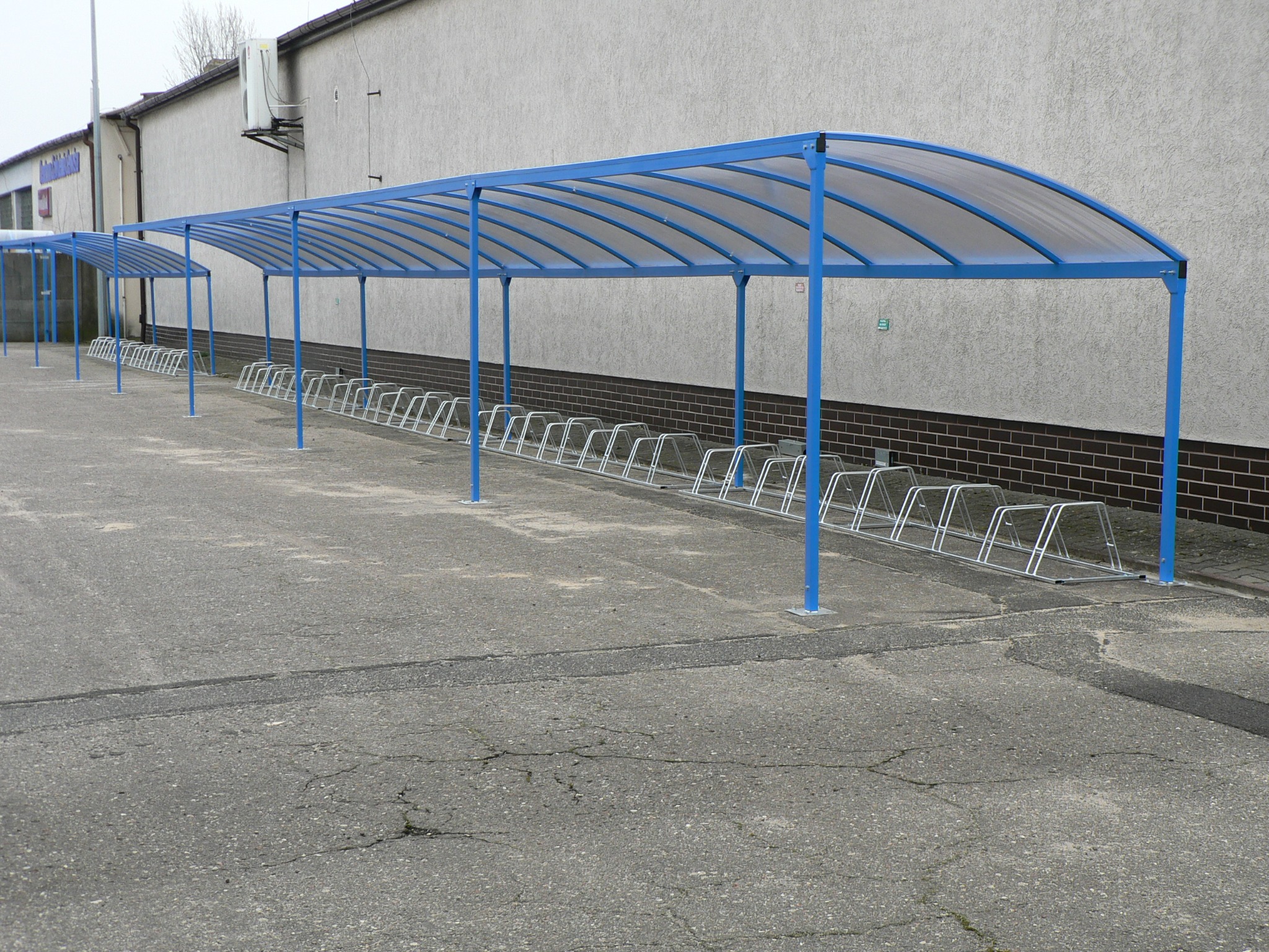 Długa, niebieska wiata rowerowa z przezroczystym zadaszeniem, stojąca na betonowym placu przed budynkiem z szarą elewacją i brązową cegłą w dolnej części, rząd metalowych stojaków na rowery pod wiatą.