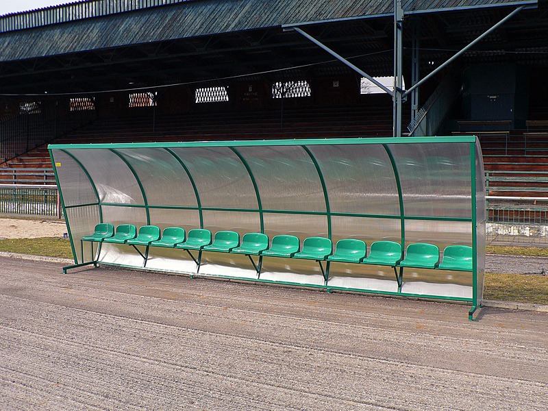 Zadaszona ławka rezerwowych, zielone plastikowe siedzenia, widok na trybuny stadionu.