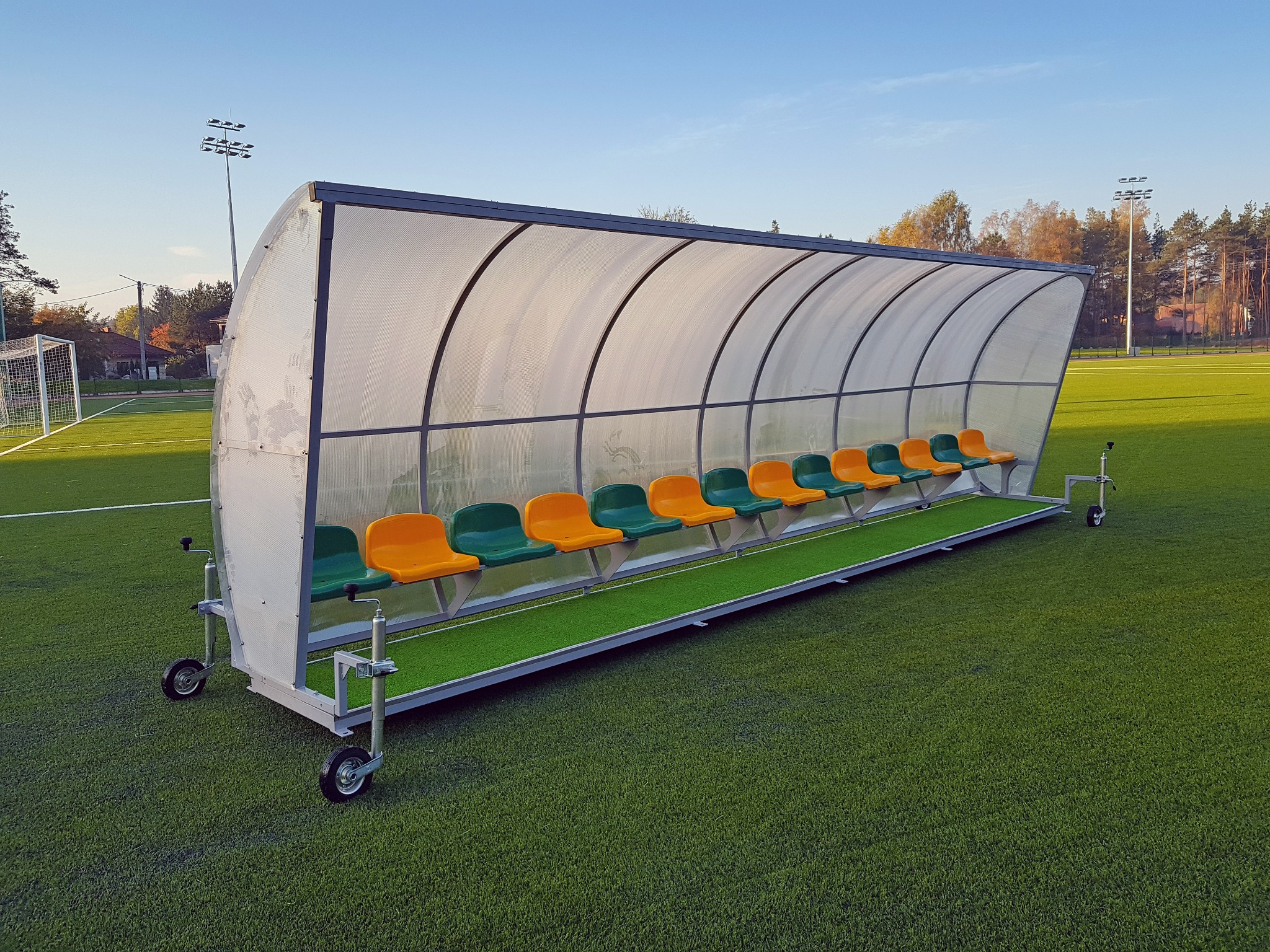 Przenośna wiata stadionowa z rzędem pomarańczowo-zielonych siedzeń, na kółkach, umieszczona na zielonym boisku, widok z boku.