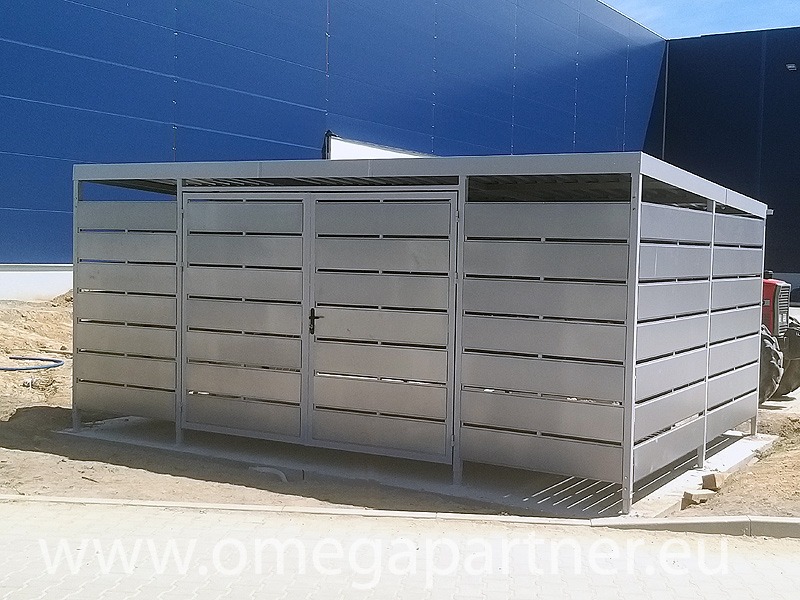 Metalowa wiata śmietnikowa z zamkniętymi drzwiami, z poziomymi szczelinami, na zewnątrz budynku z niebieską elewacją.