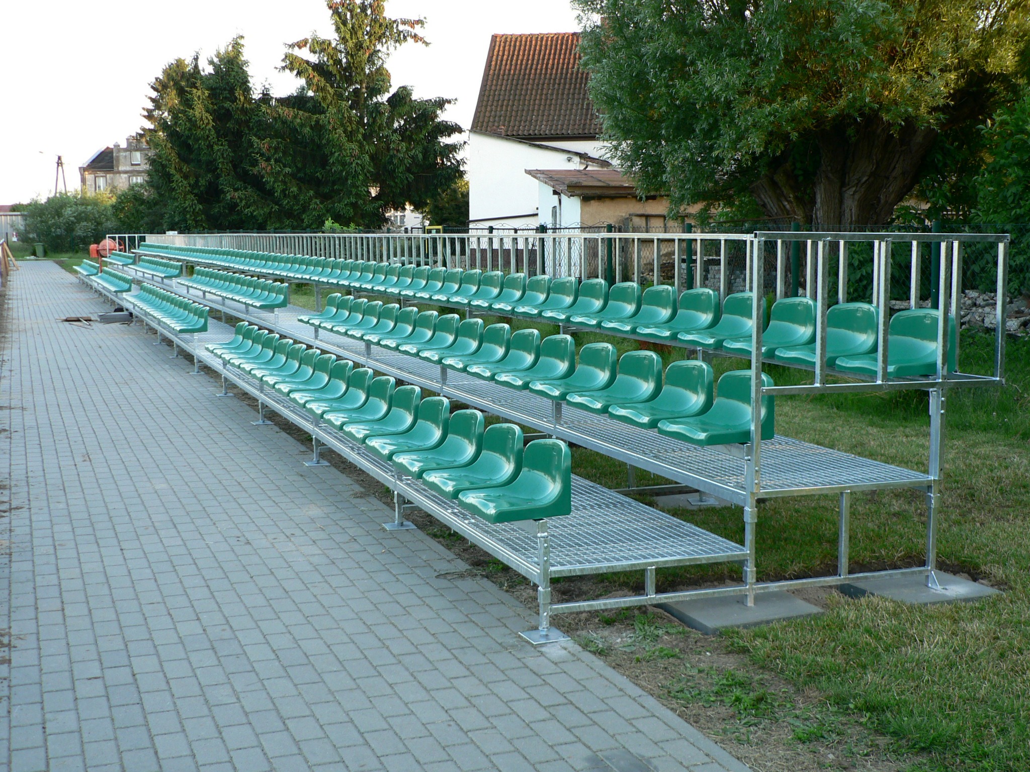 Zewnętrzna, modułowa trybuna stadionowa z zielonymi siedziskami, wykonana ze stali ocynkowanej, ustawiona na kostce brukowej i trawie.