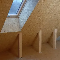 Surowe wnętrze poddasza z oknem dachowym i drewnianą konstrukcją nośną z płyty OSB, widoczne pionowe słupki podtrzymujące