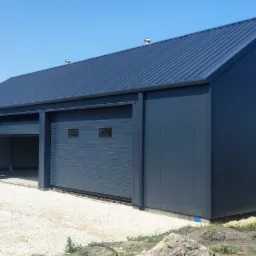 Nowoczesna hala magazynowa z szarej blachy trapezowej z trzema bramami segmentowymi, częściowo otwartymi, na żwirowym podłożu, wentylatory na dachu.