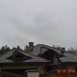 Dom w trakcie budowy z ciemnoszarą dachówką, widoczne kominy z cegły i fragment drewnianego ogrodzenia na pierwszym planie, pochmurne niebo w tle.