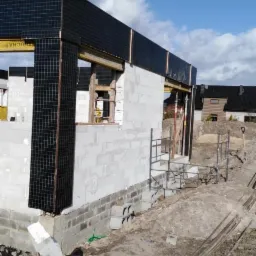 Surowy stan domu murowanego z betonu komórkowego, widoczne czarne panele elewacyjne w trakcie montażu, rusztowanie i teren budowy.