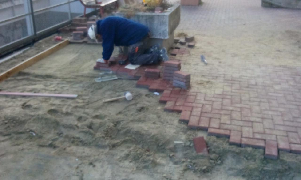 Układanie czerwonej kostki brukowej na piasku, widoczny pracownik w kasku i niebieskiej bluzie, narzędzia i częściowo ułożona nawierzchnia.