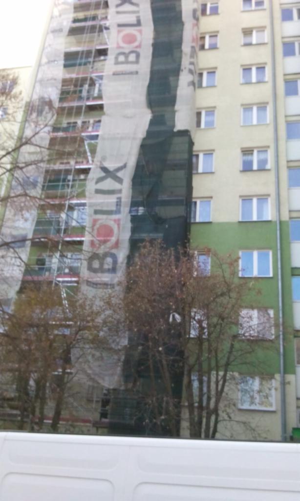 Wielopiętrowy budynek w trakcie prac elewacyjnych, zakryty siatką z reklamą marki Bolix, widoczne rusztowania i drzewa na pierwszym planie.
