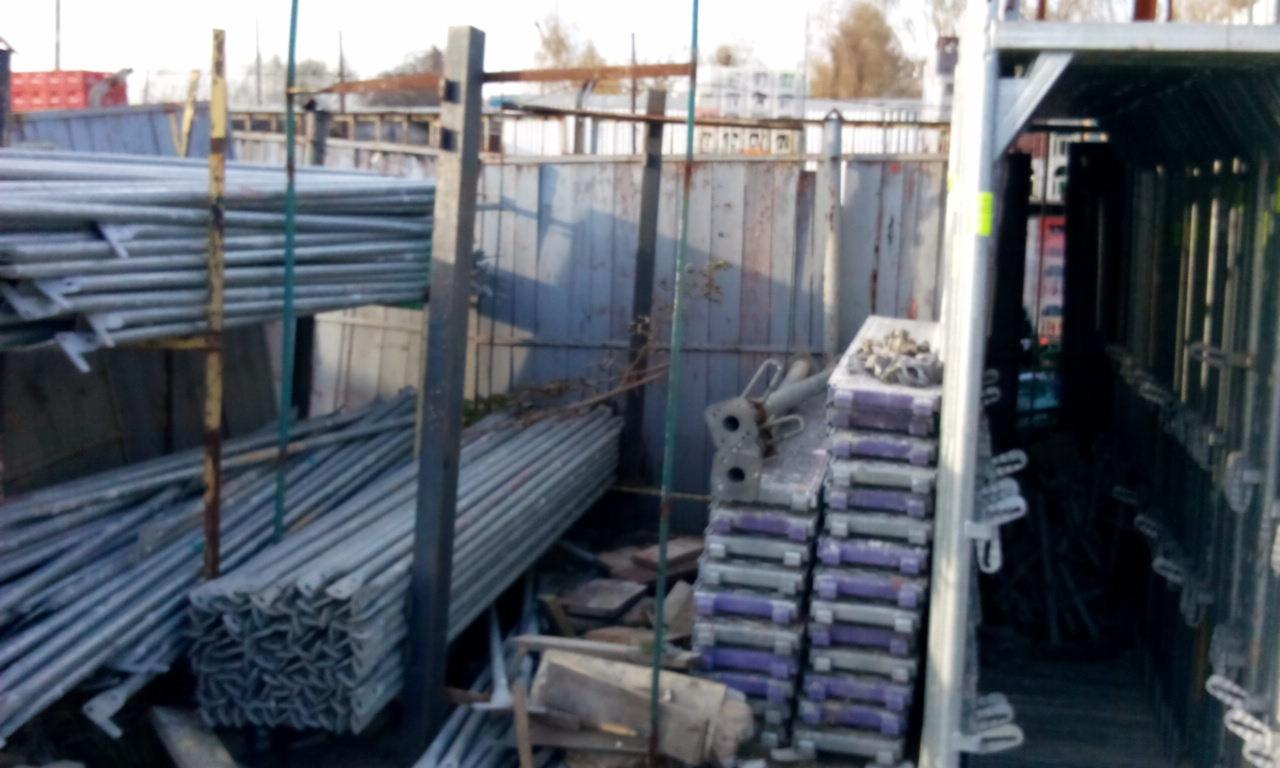 Składowisko metalowych rur, podpór budowlanych i elementów rusztowań na tle blaszanej ściany i konstrukcji metalowych.