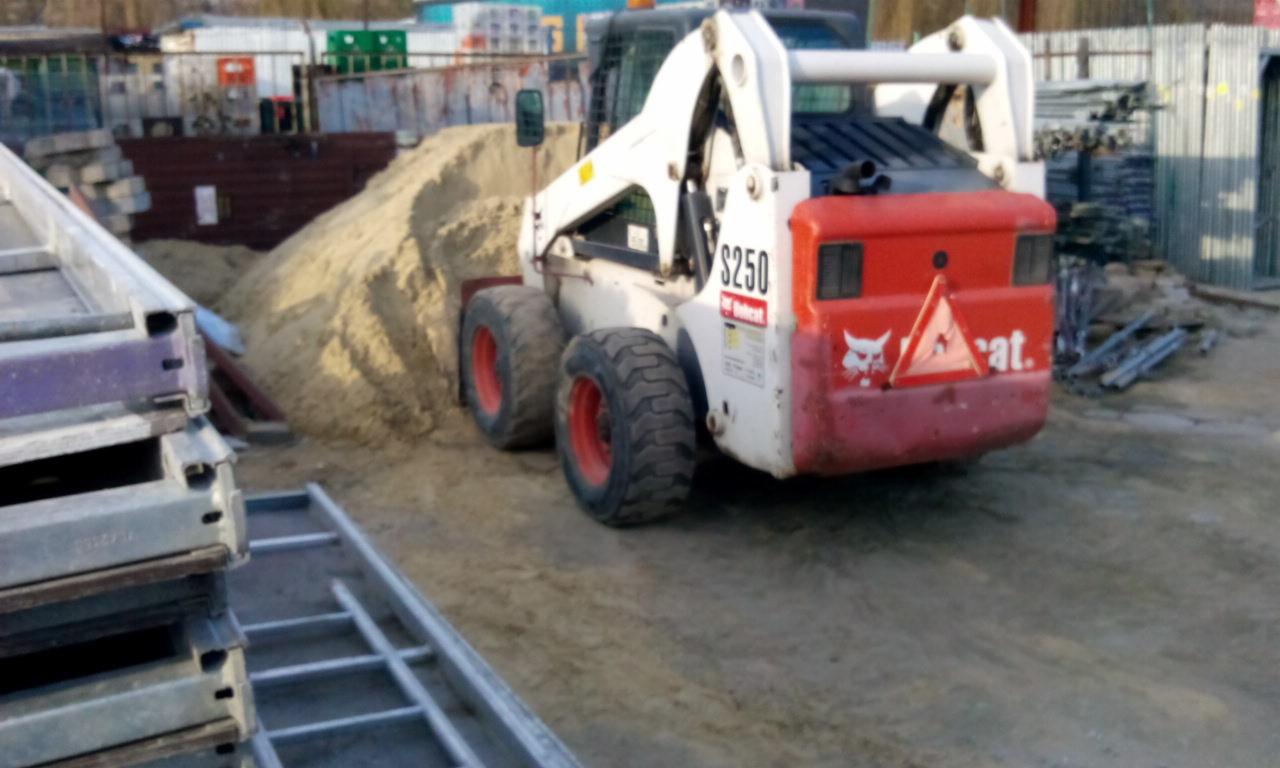 Kompaktowa ładowarka Bobcat S250 z czerwoną tylną częścią i stertą piasku na placu budowy, widoczne rusztowania i inne materiały budowlane w tle.