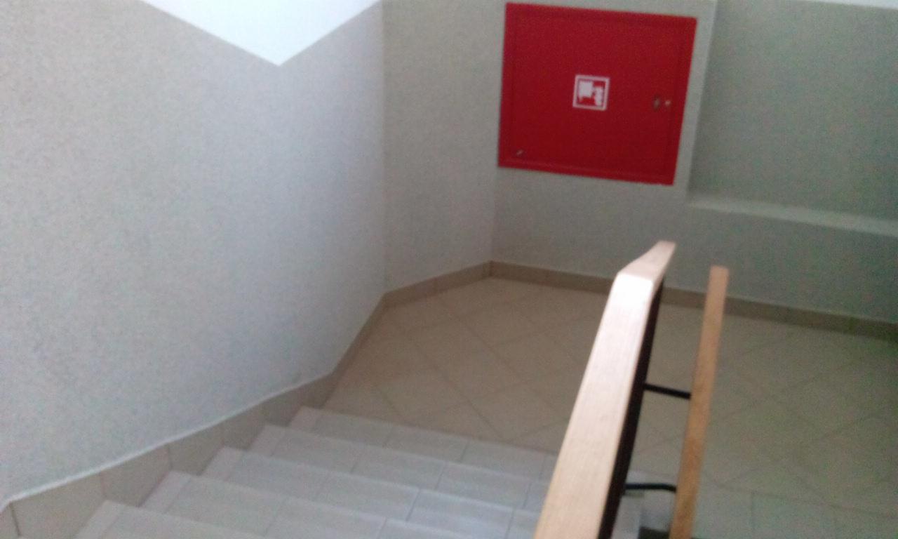 Wnętrze klatki schodowej z beżowymi płytkami na podłodze i schodach, jasnymi ścianami oraz czerwoną szafką hydrantową na ścianie.
