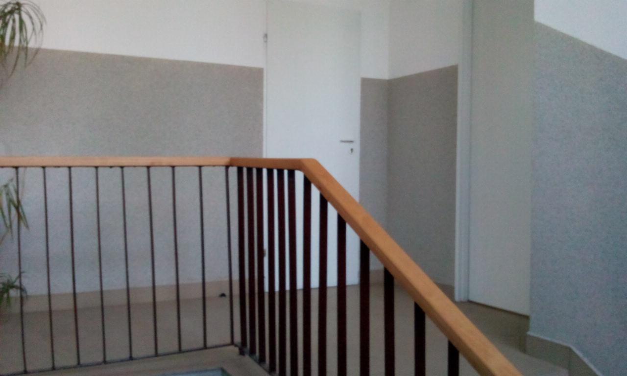 Klatka schodowa wewnątrz budynku z drewnianą balustradą i metalowymi pionowymi prętami, widok na ścianę z szarą fakturą i białe drzwi.