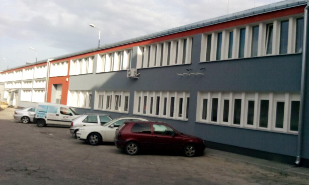 Dwupiętrowy budynek biurowy z szarą elewacją, białymi oknami i czerwonym akcentem na dachu, zaparkowane samochody przed budynkiem.