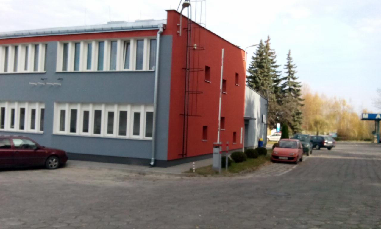 Dwukondygnacyjny budynek biurowy z elewacją pomalowaną na szaro i czerwono, z licznymi oknami, zewnętrzną metalową drabinką na czerwonej ścianie, widoczny z zewnątrz na parkingu z samochodami...