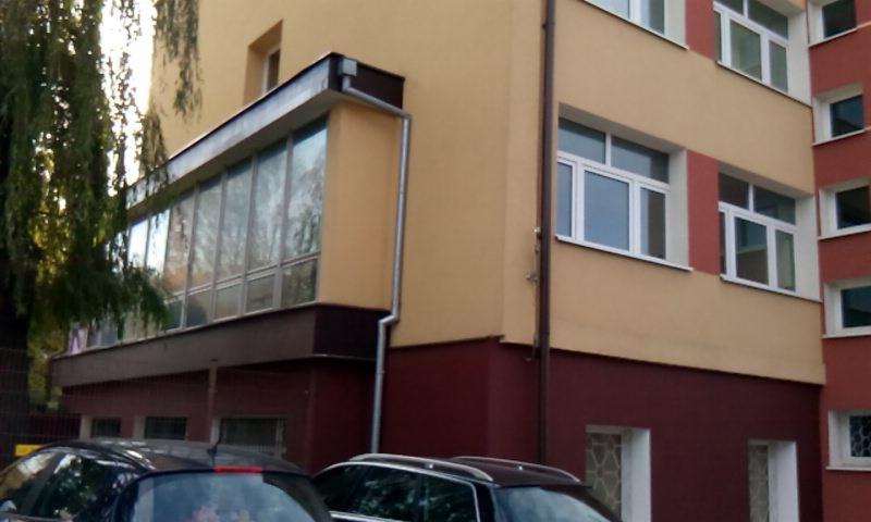 Budynek wielorodzinny z zabudowanym, przeszklonym balkonem na pierwszym piętrze, widoczne zaparkowane samochody i fragment drzewa.