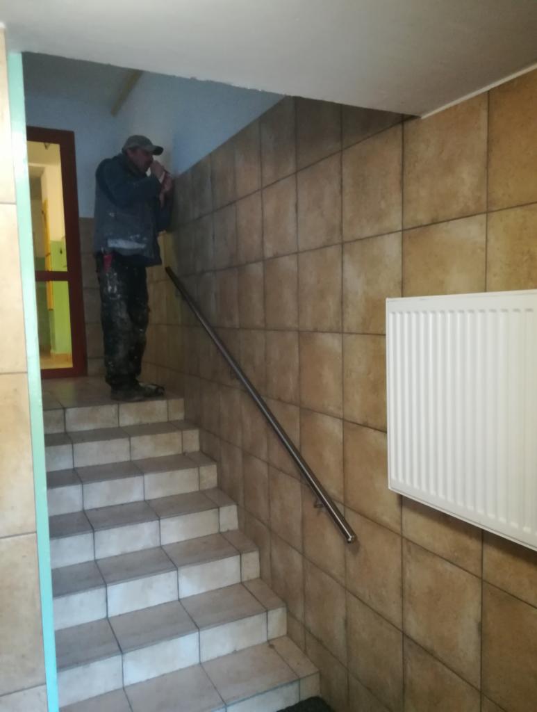 Pracownik w czapce, stojący na schodach w klatce schodowej wykończonej brązowymi kaflami, poprawia coś na ścianie w pobliżu sufitu. Widoczny grzejnik na ścianie i metalowa poręcz.