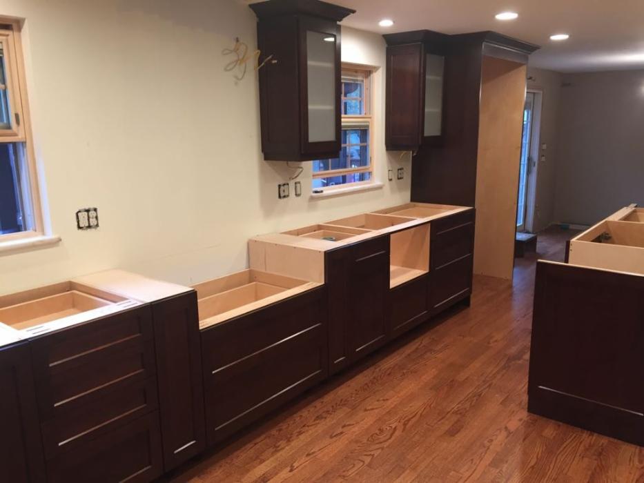 Montaż ciemnobrązowych szafek kuchennych z drewnianymi blatami w trakcie instalacji, widoczne niedokończone podłączenia elektryczne i drewniana podłoga.