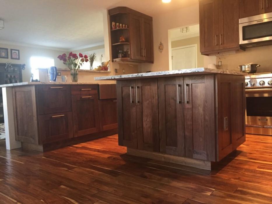 Rustykalna kuchnia z drewnianymi szafkami w odcieniach brązu, wyspą kuchenną z blatem z jasnego kamienia i podłogą z ciemnego drewna. W tle widoczny piekarnik i inne elementy wyposażenia kuchennego.