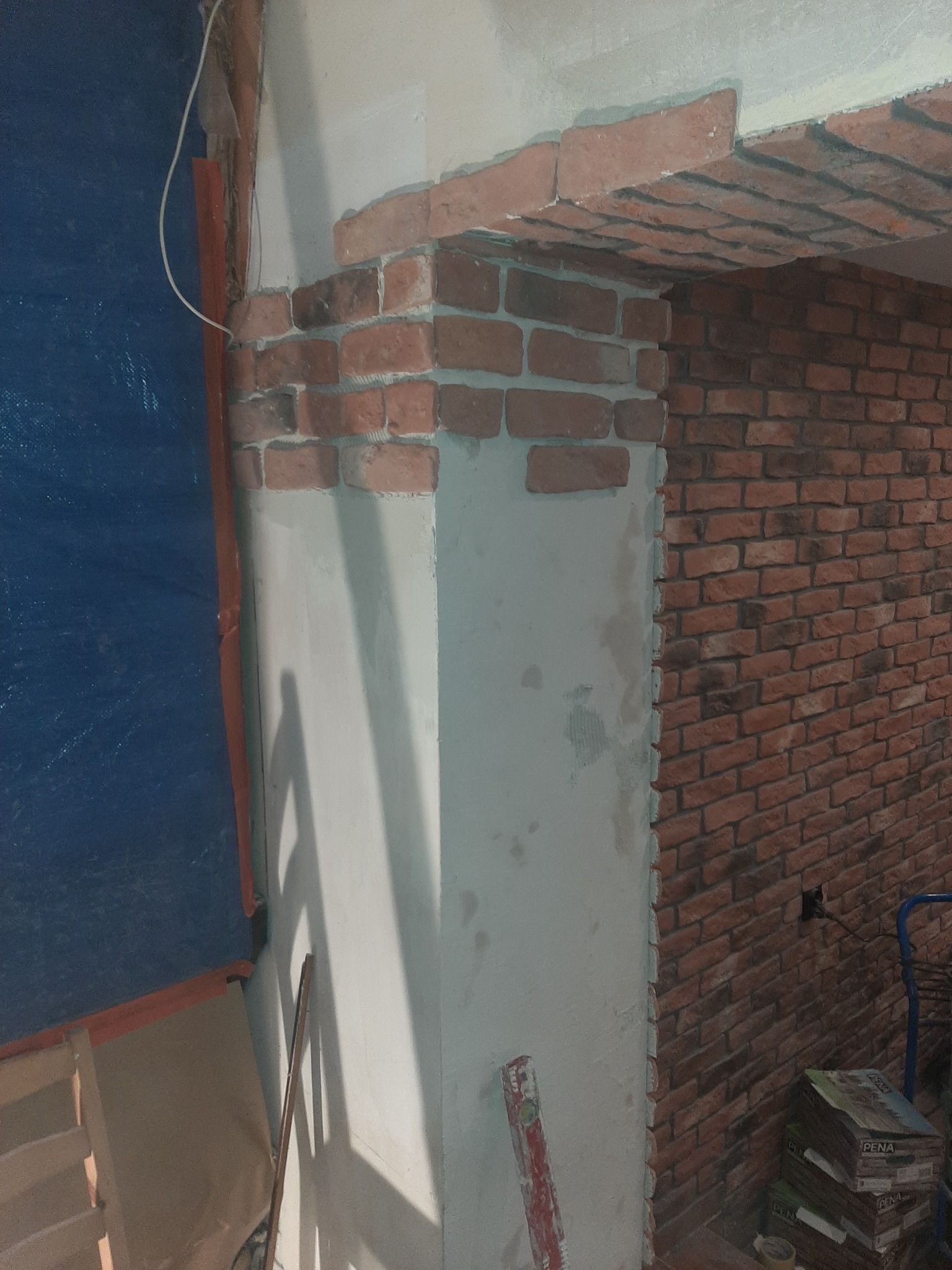 Fragment wnętrza w trakcie wykańczania z dekoracyjną cegłą, widoczna kolumna w trakcie obróbki, częściowo pokryta gładzią, oraz fragment ściany obłożonej cegłą, całość zabezpieczona folią malarską.