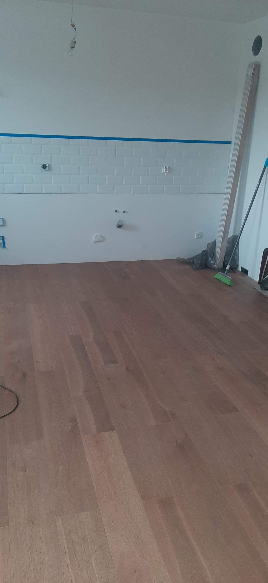 Wnętrze w trakcie prac wykończeniowych: ułożony parkiet w odcieniach brązu, ściana z białymi płytkami typu metro zabezpieczona niebieską taśmą malarską, widoczne gniazdka elektryczne i rury,...