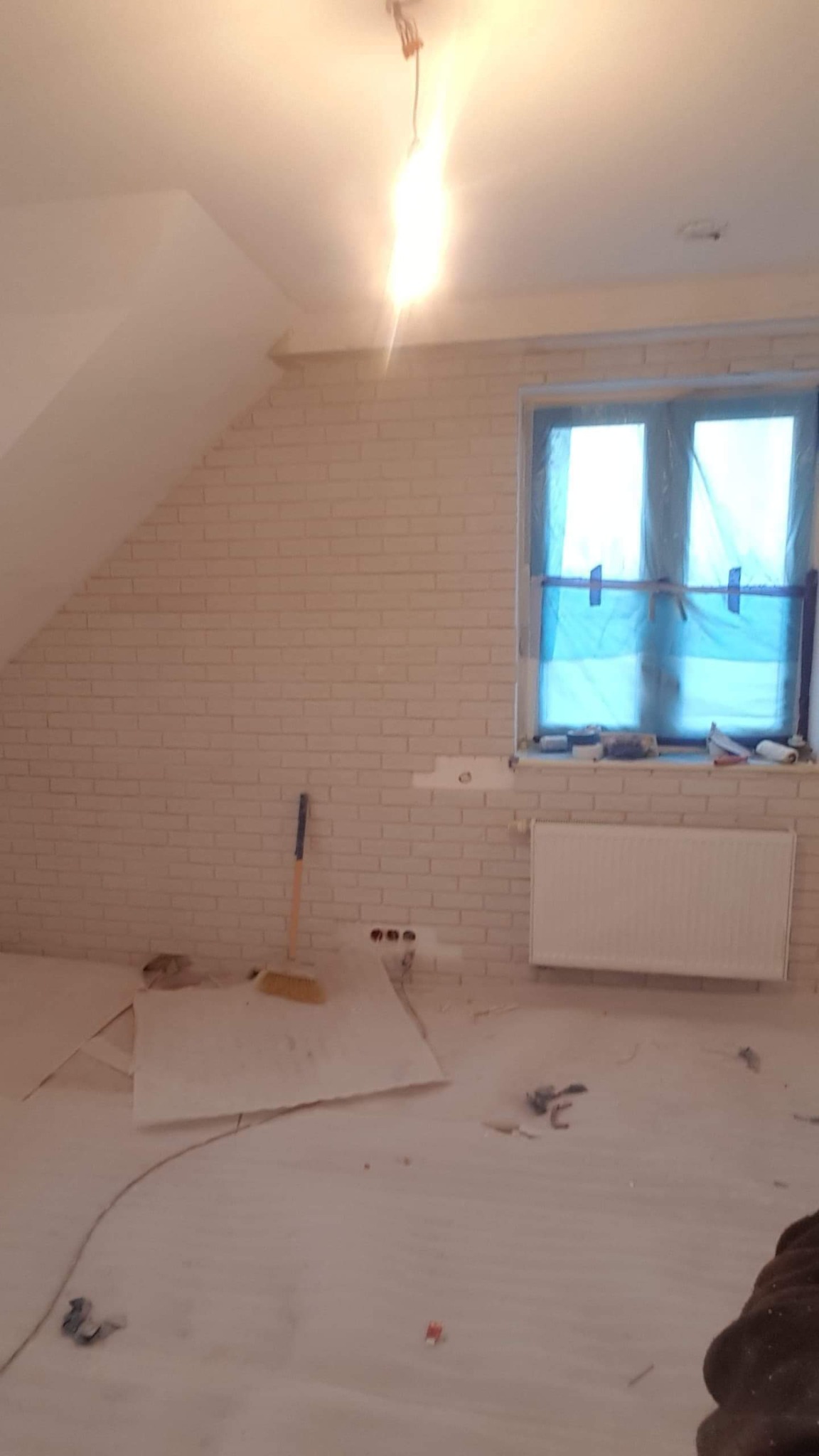 Pokój na poddaszu w trakcie wykańczania, ściana wyłożona białą cegłą, okno zabezpieczone folią malarską, widoczne gniazdka elektryczne, grzejnik i narzędzia malarskie.