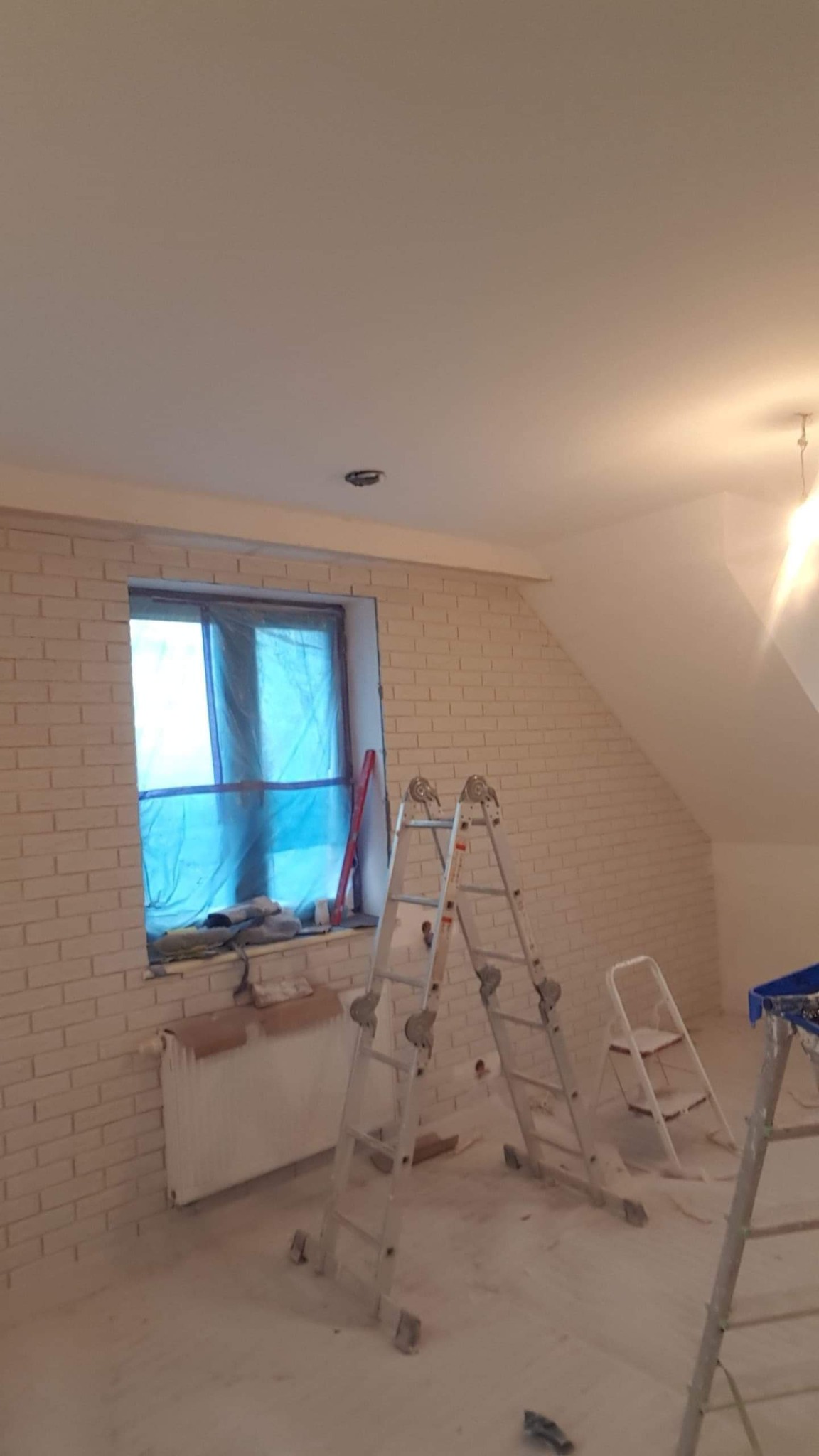 Pokój na poddaszu po malowaniu ścian imitujących cegłę, okno zabezpieczone folią, widoczne drabiny malarskie i pomalowany grzejnik.