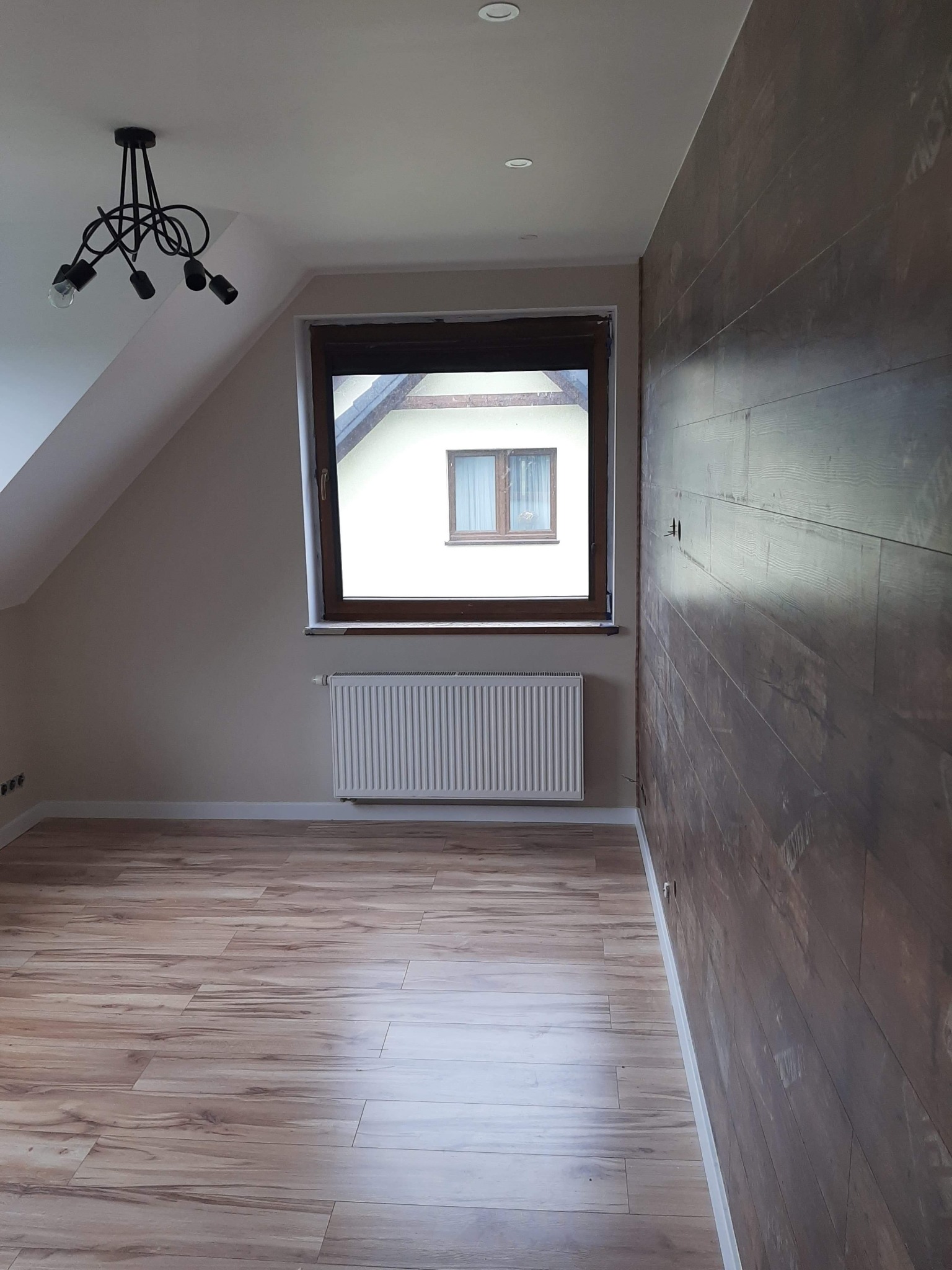Poddasze po remoncie: panele podłogowe imitujące drewno, ściana wykończona panelami ściennymi w odcieniach brązu, biały grzejnik pod oknem, czarna lampa sufitowa.