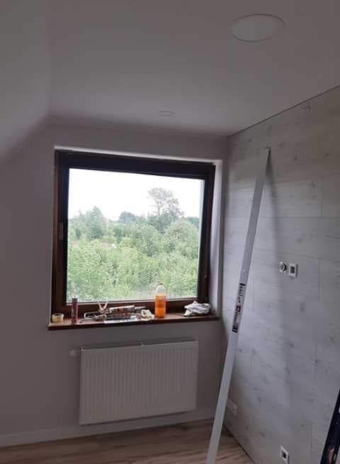 Pokój w trakcie remontu: panele ścienne imitujące drewno, okno z widokiem na zieleń, narzędzia na parapecie, poziomica oparta o ścianę.