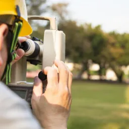 Osoba w kasku obsługuje niwelator na zielonym terenie, w tle druga osoba z łatą niwelacyjną.