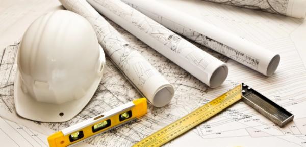 Plan sytuacyjny z białym kaskiem, żółtą poziomicą, kątownikiem i zwiniętymi projektami technicznymi.