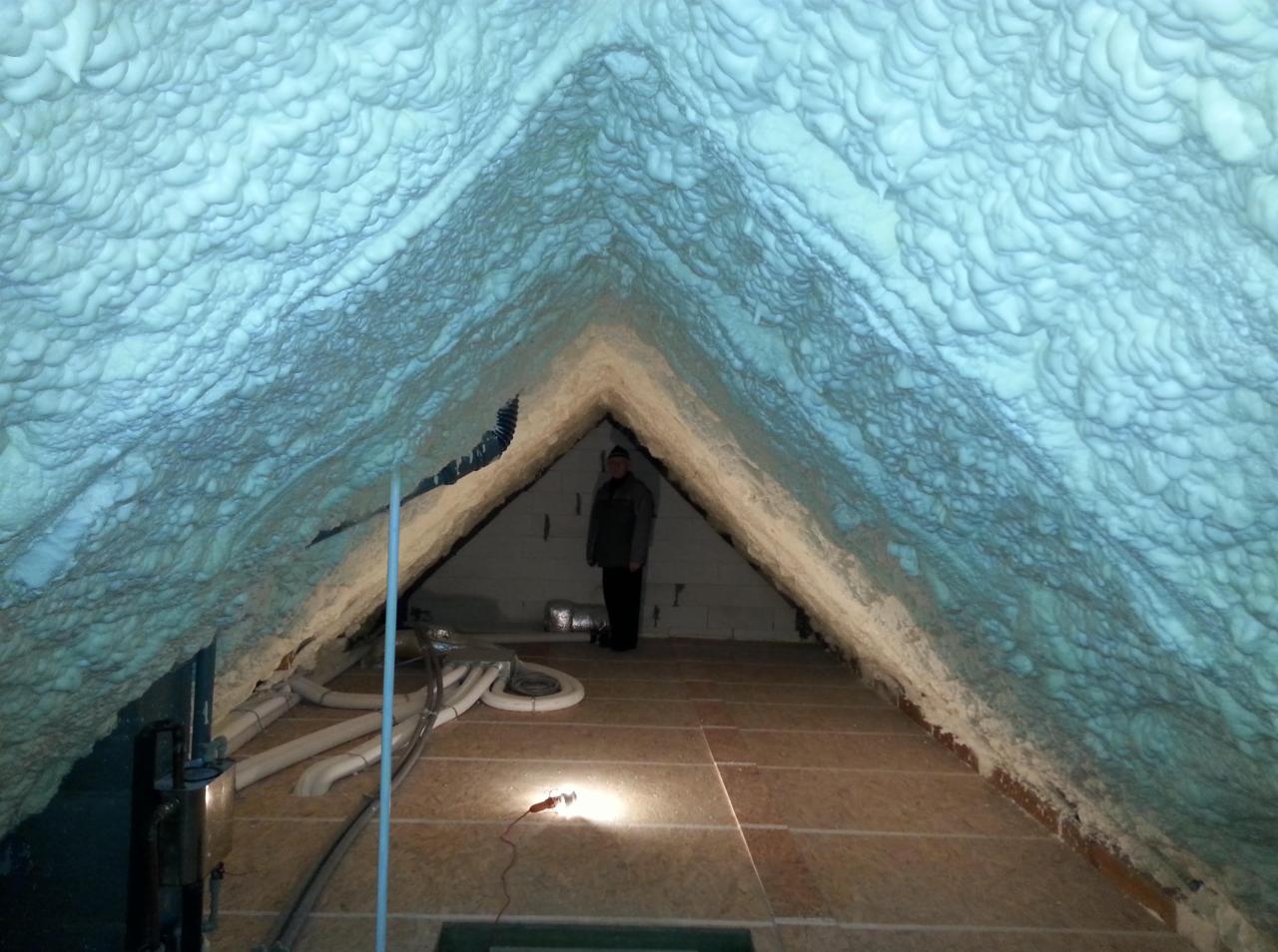 Poddasze ocieplone pianką z widocznymi przewodami i pracownikiem w tle, oświetlone lampą halogenową leżącą na podłodze.