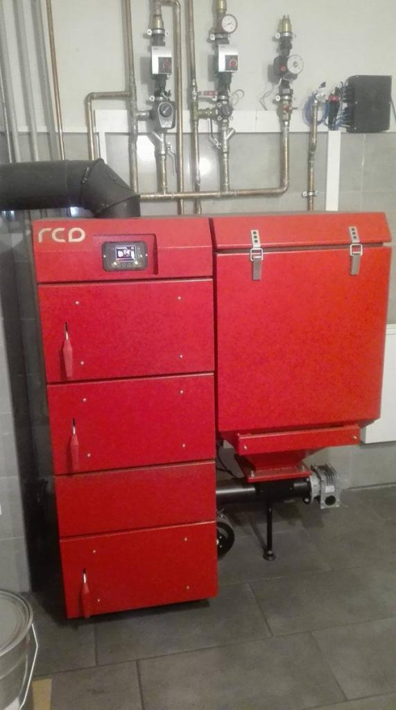 Czerwony, nowoczesny piec na paliwo stałe z podajnikiem, zainstalowany w kotłowni z widocznymi rurami i armaturą na ścianie.