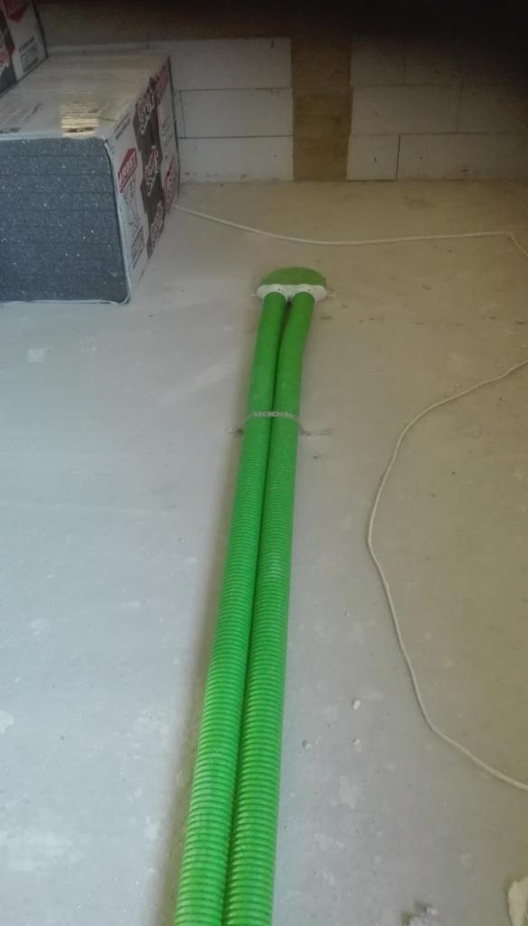 Dwa zielone, karbowane przewody wentylacyjne wychodzące ze ściany, zabezpieczone opaską zaciskową, widoczne materiały budowlane w tle.