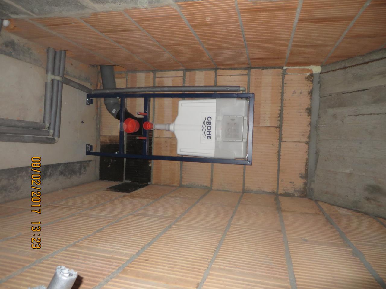 Stelaż podtynkowy Grohe zamontowany na ceglanej ścianie, widoczne rury instalacyjne i fragment posadzki z cegły.