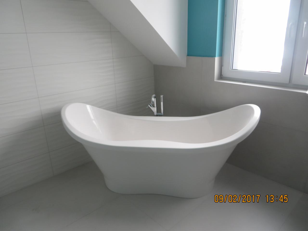 Wolnostojąca wanna w kształcie łódki z chromowaną baterią, umieszczona w łazience z białymi płytkami o falistej strukturze i turkusowym akcentem kolorystycznym przy oknie.