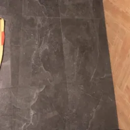 Wykończona podłoga prysznica z ciemnoszarych płytek imitujących kamień, z odpływem liniowym, sąsiadująca z drewnopodobną podłogą w łazience.