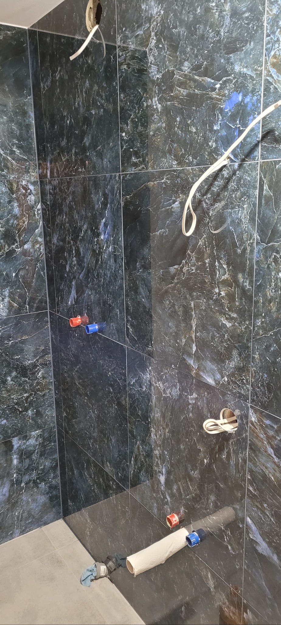 Ściana łazienki wyłożona ciemnymi płytkami imitującymi marmur, widoczne wystające przewody elektryczne i rury wodne, etap prac wykończeniowych.