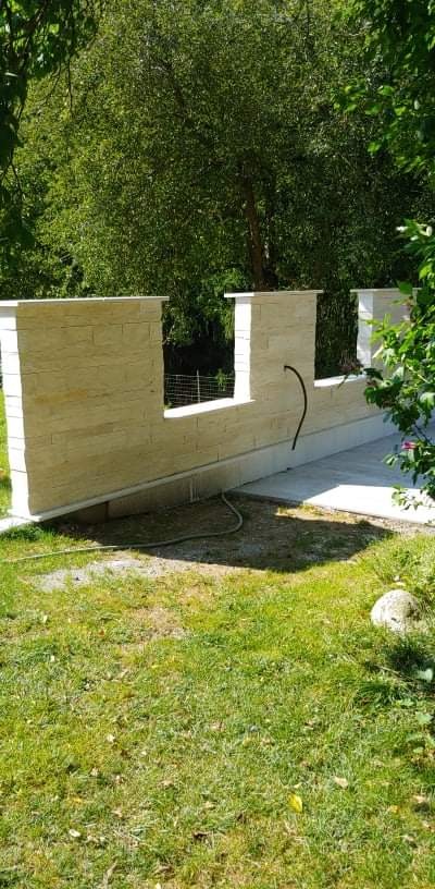 Murowana ściana z piaskowca w ogrodzie, z widoczną instalacją, na tle zieleni.