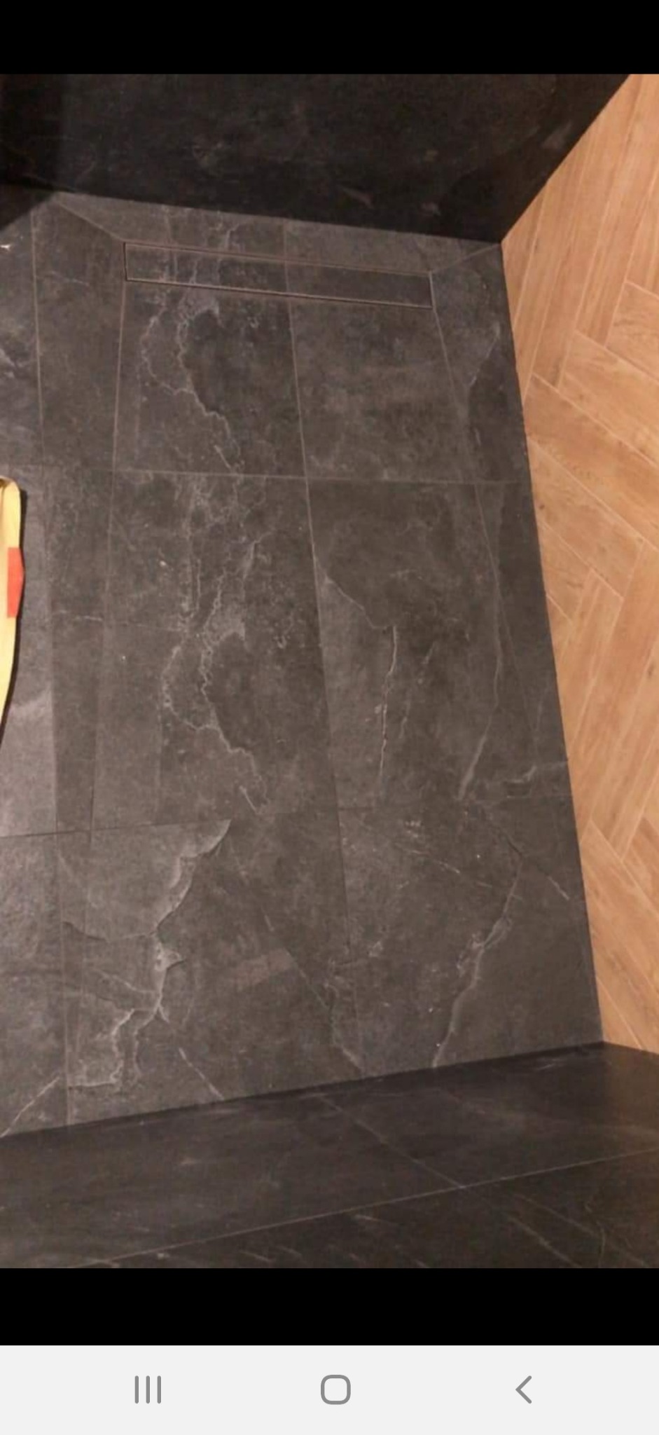 Wykończona podłoga prysznica z ciemnoszarych płytek imitujących kamień, z odpływem liniowym, sąsiadująca z drewnopodobną podłogą w łazience.