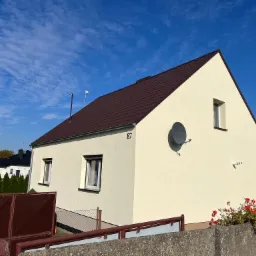 Dom jednorodzinny z brązowym dachem z blachodachówki, jasną elewacją i numerem 87, widziany zza betonowego murku porośniętego czerwonymi kwiatami.