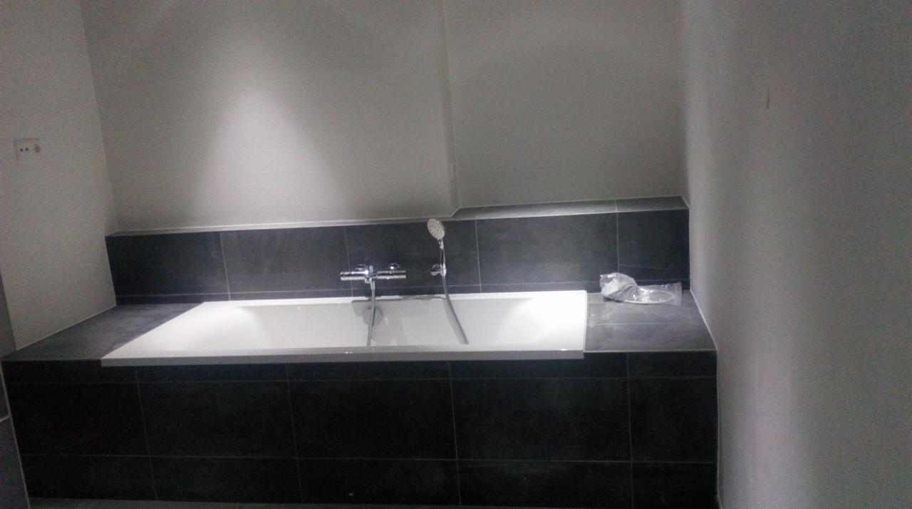 Nowoczesna, prostokątna wanna obudowana ciemnymi płytkami, bateria i słuchawka prysznicowa zamontowane na krawędzi wanny, widoczne niedoskonałości wykończenia na ścianie i folia ochronna na płytkach.