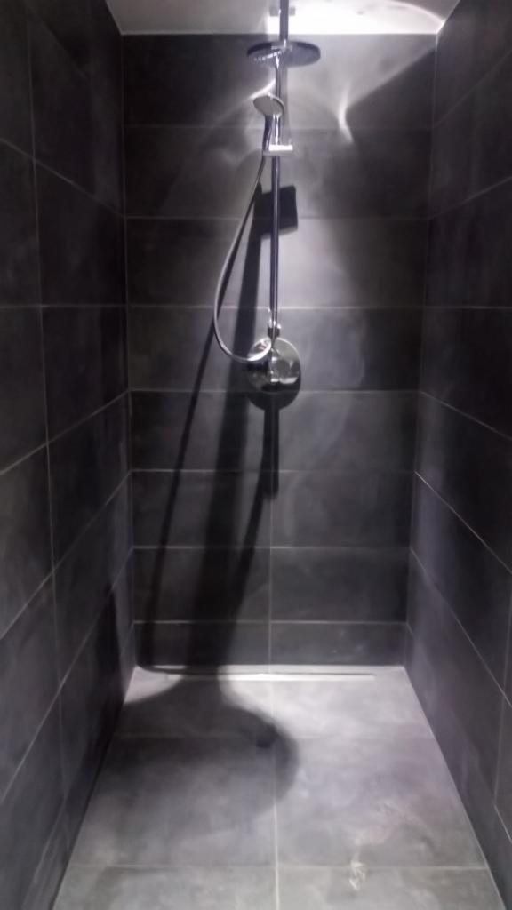 Nowoczesna kabina prysznicowa z ciemnoszarymi płytkami, deszczownicą i odpływem liniowym w podłodze, oświetlona górnym światłem tworzącym refleksy na ścianach.