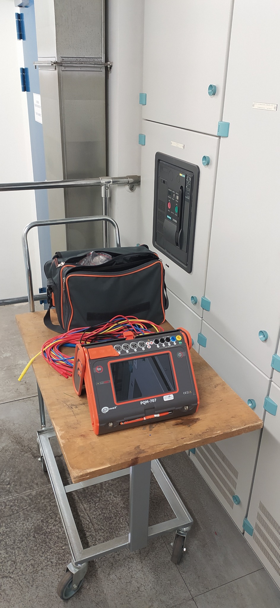 Miernik parametrów elektrycznych Sonel PQM-707 z przewodami pomiarowymi i torbą narzędziową na metalowym wózku na tle rozdzielnicy elektrycznej.