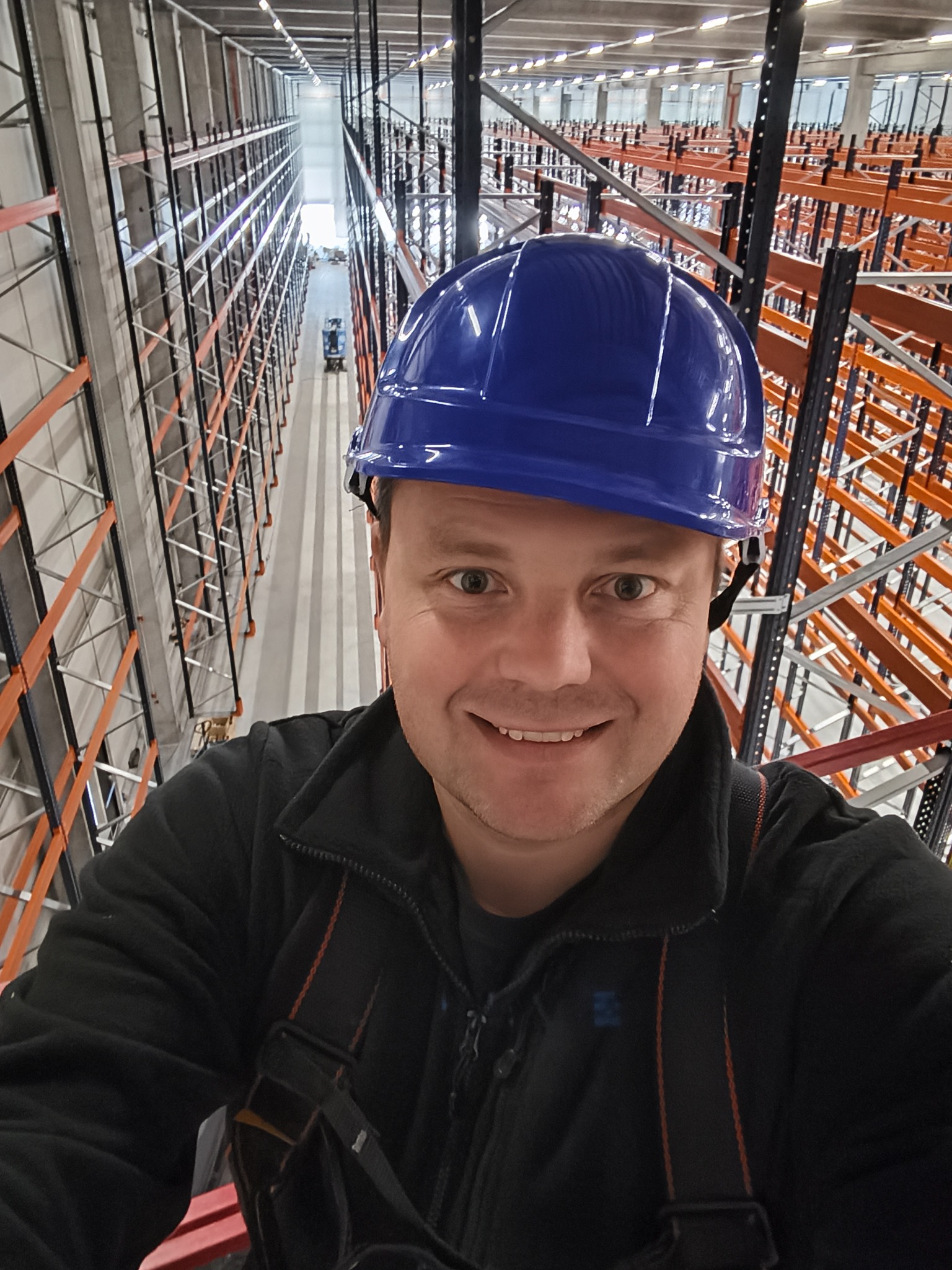 Uśmiechnięty mężczyzna w niebieskim kasku na tle wysokich regałów magazynowych. Widok z góry na wózek widłowy w oddali. Oświetlenie sufitowe.