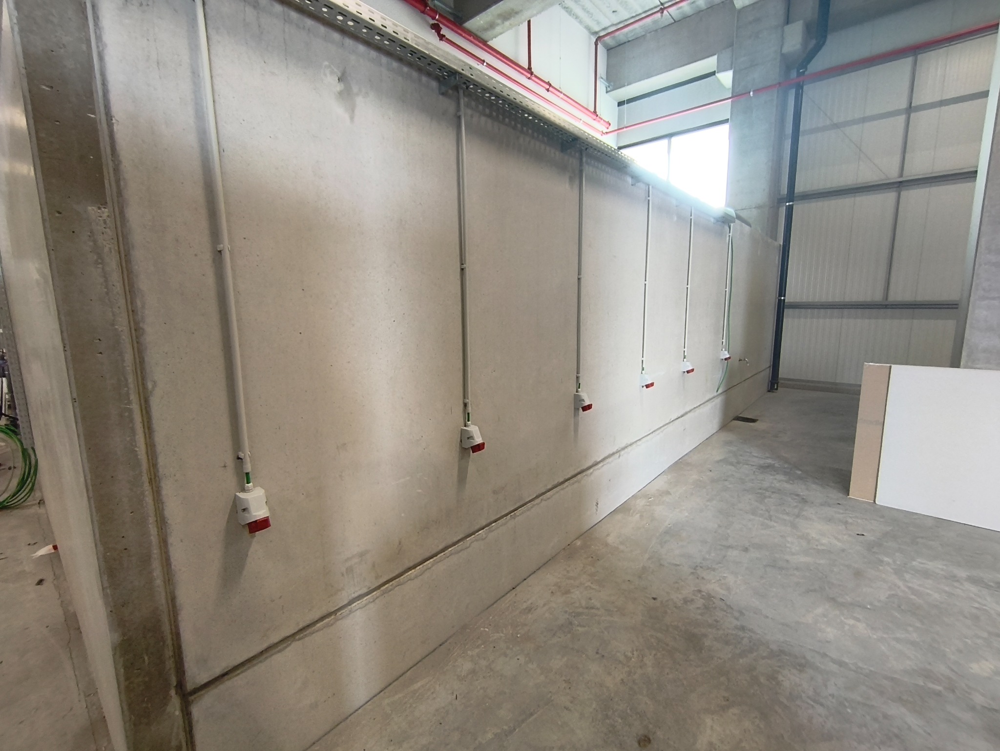 Szereg gniazd elektrycznych z czerwonymi wtykami, zamontowanych na betonowej ścianie w hali przemysłowej. Widoczne koryta kablowe pod sufitem i betonowa podłoga.