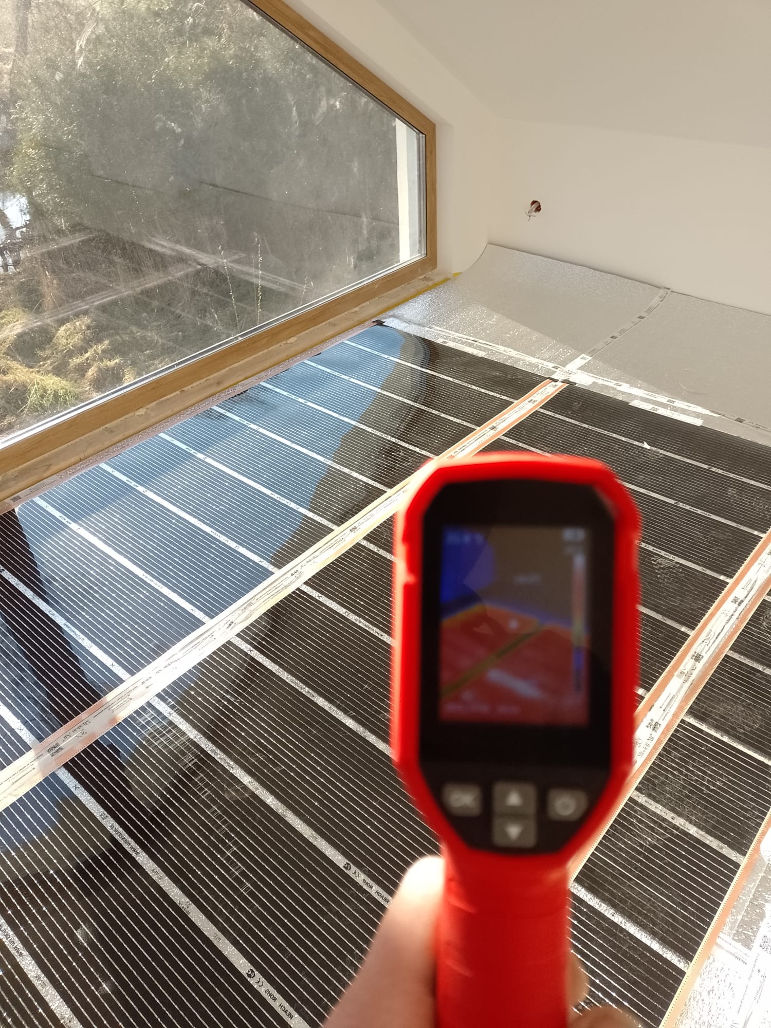 Termowizja maty grzewczej pod oknem, na izolacji. Czerwony termometr w dłoni, widok z góry na czarne pasy maty i okno z widokiem na zewnątrz.