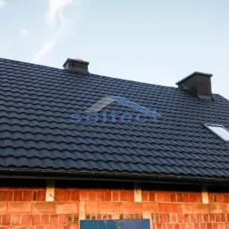 Czarny dach z blachy na budynku w trakcie budowy, widoczne kominy i okno dachowe, ściany z czerwonej cegły.