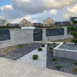 Nowoczesne ogrodzenie panelowe z podmurówką z bloczków betonowych, brama wejściowa i ścieżka z białego grysu na tle domów jednorodzinnych.